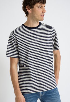 Mies, jolla on tummansininen ja valkoinen vaakaraitainen lyhythihainen t-paita, jossa on tummansininen kaulus, katsoo sivulle, siniset farkut, neutraali tausta.