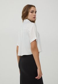 Camicetta bianca corta con maniche corte e colletto, caratterizzata da una piega sul retro. Indossata con una gonna nera, tessuto liscio.