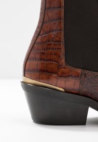 Gros plan sur une botte à chevilles marron embossée crocodile, avec un panneau élastique noir et un petit talon en bois foncé agrémenté d'un accent métallique.