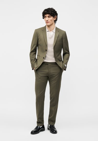 Άνδρας που φοράει ελαιόχρωμο κοστούμι με ανοιχτό μπεζ πλεκτό πόλο πουκάμισο και μαύρα επίσημα παπούτσια, στέκεται με τα χέρια στις τσέπες σε μονόχρωμο φόντο.