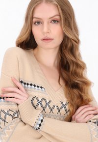 Beżowy sweter dzianinowy z dekoltem w kształcie litery V, z wzorzystymi akcentami w niebieskie i czarne geometryczne wzory na rękawach. Miękka tekstura, luźny krój.
