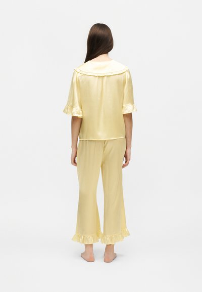 Femme aux longs cheveux bruns, debout pieds nus de dos, portant un pyjama en satin jaune pâle avec un col, des manches et des ourlets de pantalon à volants.