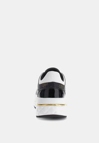 Zapatilla en blanco y negro con un talón negro brillante, acento dorado en la suela y tejido texturizado. Presenta un logo en la parte trasera y un cuello acolchado.