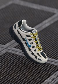 Balti un melni Nike P-6000 sporta apavi ar zilu swoosh un dzeltenām auklām, novietoti uz metāla restes virsmas.