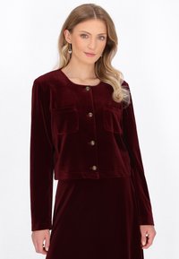 Bordeaux fluwelen cropped jacket met lange mouwen, twee voorzakken en gouden knoopsluitingen. Zachte textuur en gladde afwerking.