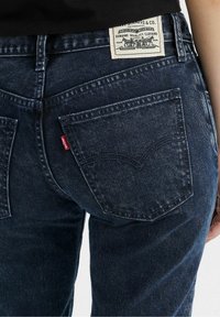 Jean en denim bleu foncé à coupe droite, avec des surpiqûres blanches contrastantes et une étiquette en cuir de marque sur la ceinture.