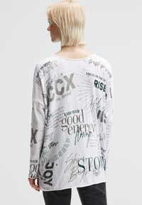 Frau mit kurzem blonden Haar, die ein weißes Langarmshirt mit schwarzem motivierendem Text und grafischen Designs trägt, steht vor einem schlichten Hintergrund.