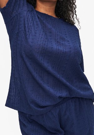 Femme portant un haut texturé bleu marine à manches courtes et un pantalon assorti, levant un bras, cadrée au niveau du cou et des hanches.
