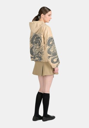 Giovane donna con i capelli legati indossa una giacca beige con cappuccio con stampa di drago nero, gonna beige, calze nere alte fino al ginocchio e scarpe nere, di profilo.