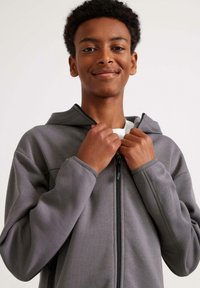 Harma zip-up collegepaita pehmeällä tekstuurilla, jossa on huppu ja säädettävä vetoketju. Malli pitää vetoketjusta molemmilla käsillä, keskittyen istuvuuteen.