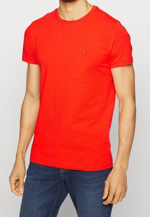 Mand iført en rød kortærmet crew neck t-shirt med lille logo på brystet og mørkeblå jeans mod en ensfarvet baggrund.