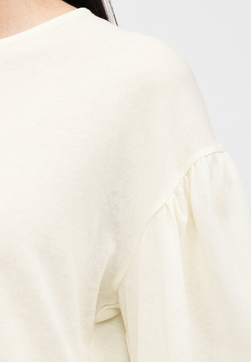 Haut de couleur crème avec un col arrondi et des manches bouffantes. Le tissu semble doux et texturé, avec un design léger et aérien.