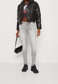 Svart kort puffjacka, grafisk crop top, ljusgrå skinny jeans och svarta sneakers; håller en liten svart handväska med texturerad yta.