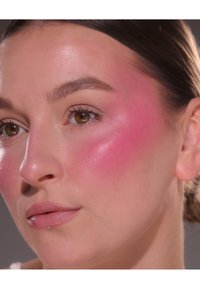Nærbilde av et ansikt med glatt hud og en glansfull finish, dyp rosa blush på kinnet og nøytral leppefarge. Brune øyne og rett brunt hår.