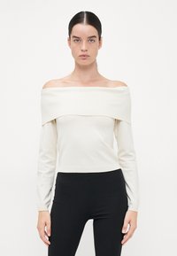 Off-shoulder langermet topp i krem ribbet stoff; tettsittende silhuett med vid halslinje som reflekterer en jevn tekstur.