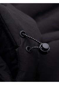 Gros plan sur un tissu noir avec deux œillets noirs et un cordon élastique noir maintenu par un bouton-pression plastique rond.