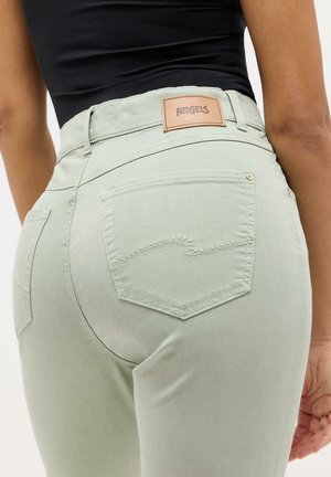 Femme portant un jean moulant vert menthe avec poches arrière et une étiquette beige "ANGELS", associé à un haut noir.