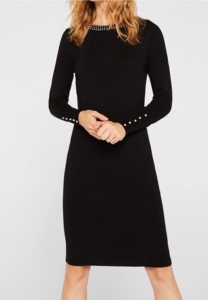 Femme portant une robe noire à manches longues jusqu'aux genoux avec des détails en perles au col et aux manches, debout les mains jointes devant elle.