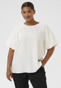Una donna con capelli scuri raccolti in un basso chignon indossa una blusa bianca a maniche corte svasate e morbide, insieme a pantaloni neri, in piedi con una mano nella tasca.