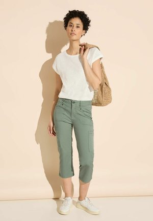 White textured short-sleeve top with green cropped pants and white sneakers. Carrying a woven tan bag, standing against a light background.