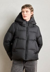Veste matelassée noire avec capuche, dotée d'une fermeture éclair à l'avant, d'un design matelassé et d'une texture douce. Portée avec un jean gris sur un fond neutre.
