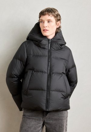 Giacca a vento nera con cappuccio, dotata di zip frontale, design trapuntato e texture morbida. Indossata con jeans grigi su uno sfondo neutro.