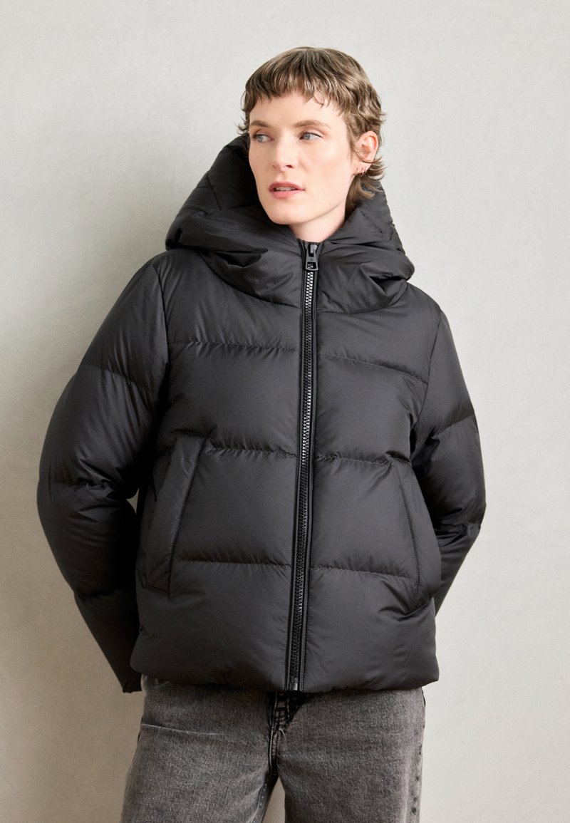 Veste matelassée noire avec capuche, dotée d'une fermeture éclair à l'avant, d'un design matelassé et d'une texture douce. Portée avec un jean gris sur un fond neutre.