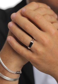 Hõberõngas musta ristkülikukujulise kiviga, kantud nimetissõrmel, kerge päevitusega sõrmede vahel. Sile pind, minimalistlik disain.