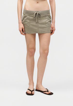 Kobieta w zielonej mini spódniczce ze sznurkiem i dużymi przednimi kieszeniami oraz czarnych klapkach, stojąca na tle jednolitego białego tła.