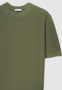 T-shirt vert olive en maille avec col rond et manches courtes, présentant une surface texturée et des finitions côtelées. Étiquette de la marque visible au niveau du col.