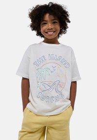 T-shirt en coton blanc avec un design brodé représentant des palmiers, un soleil et des vagues, associé à un pantalon de survêtement jaune clair.