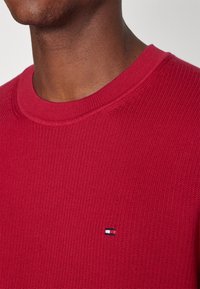 Suéter de punto rojo con cuello redondo, presenta textura de canalé y un pequeño logo en el pecho. La tela parece suave y casual.