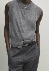Gray sleeveless cardigan with a high collar, featuring a button front and ribbed texture, paired with gray trousers and a black belt.