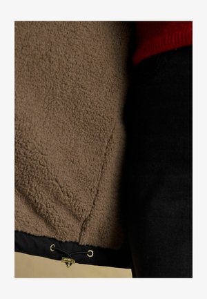 Braune Jacke mit Fleece-Futter und schwarzer Außenseite, mit einem Kordelzugverschluss am unteren Ende. Weicher, strukturierter Stoff sichtbar.