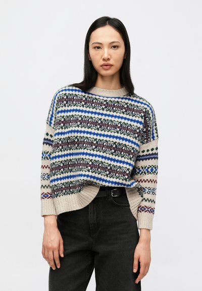 Femme aux longs cheveux foncés portant un pull ample beige, bleu, vert et bordeaux à motifs et un jean noir avec une ceinture noire.