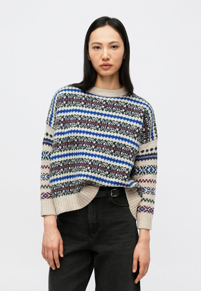 Woman with long dark hair wearing a loose beige, blue, green, and maroon patterned sweater and black jeans with a black belt.