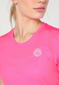 Světle růžové sportovní tričko s kulatým výstřihem, síťovanou texturou a bílým kruhovým logem na levé straně hrudi.