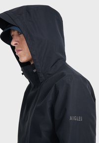 Veste imperméable navy avec capuche, dotée d'un col haut, d'une fermeture avant et du logo de la marque "AIGLE 1853" sur la manche.