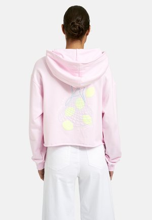 Frau mit dunklen Haaren im Dutt, trägt einen kurz geschnittenen hellrosa Hoodie mit Zitronenmotiv und weiße Hose, steht mit dem Rücken zur Kamera vor schlichter Hintergrund.