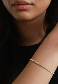 Złota bransoletka z rzędem naprzemiennych przezroczystych i niebieskich kamieni, smukły design, gładka tekstura, noszona na opalonej nadgarstku na ciemnym tle.