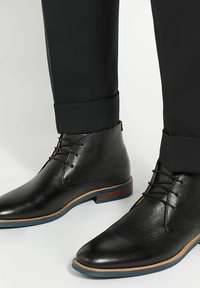 Bottines en cuir noir avec une texture lisse et un bout pointu, présentant des accents marron et une semelle extérieure bleue, associées à un pantalon sombre.