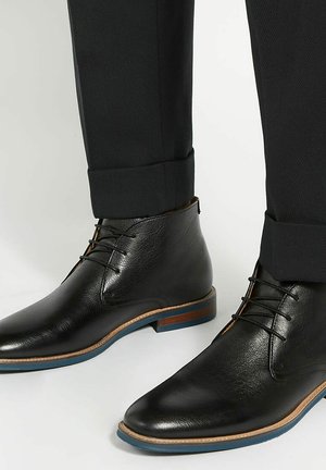 Bottines en cuir noir avec une texture lisse et un bout pointu, présentant des accents marron et une semelle extérieure bleue, associées à un pantalon sombre.