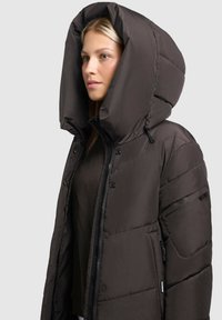 Ung kvinna i en mörkbrun vadderad vinterjacka med stor huva, som står mot en enfärgad ljus bakgrund.