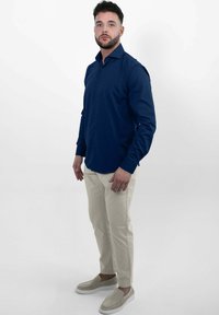 Marineblauw overhemd met lange mouwen, gecombineerd met lichtbeige tapered broek en beige instappers, gedragen door een staande mannelijke model.