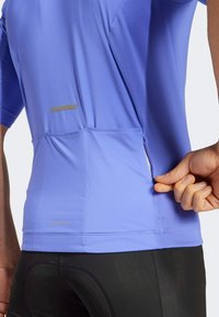Top sportivo di colore viola chiaro con maniche corte, tessuto testurizzato, tasca laterale con zip e dettaglio riflettente sul retro.