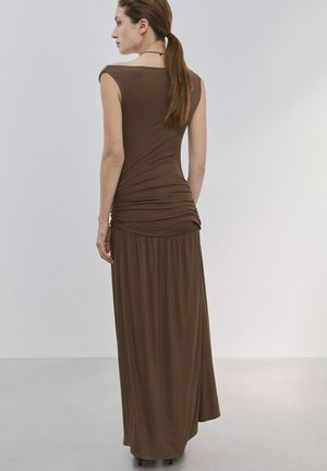 Mujer con coleta marrón, vistiendo un vestido largo marrón sin mangas con fruncidos, de pie y de espaldas sobre un fondo liso.