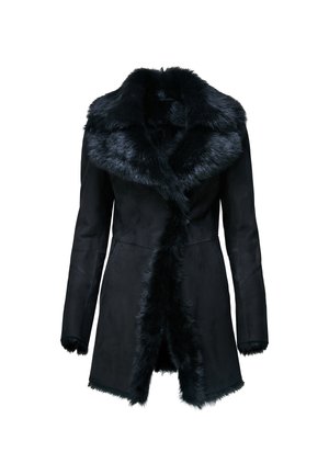 Femme portant un manteau noir bordé de fourrure, chemise marron, pantalon en cuir noir, talons noirs, tenant un sac à main en fourrure à motif léopard, debout sur un fond neutre.