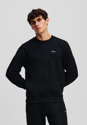 Sort sweatshirt lavet af blødt stof, med rund halsudskæring og lange ærmer. Indeholder en lille, rektangulær logo-patch på brystet.