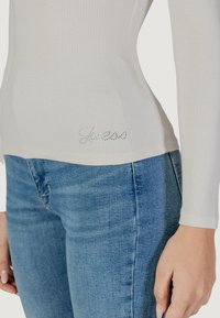 Top a manica lunga aderente a costine in beige chiaro con una finitura testurizzata, caratterizzato da un logo "Guess" argentato impreziosito all'orlo. Abbinato a jeans blu.