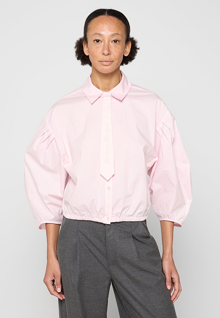 Blusa rosa con un cuello estructurado y un detalle de lazo. Presenta mangas abullonadas y recogidas, y un dobladillo cortado, hecha de tela ligera.